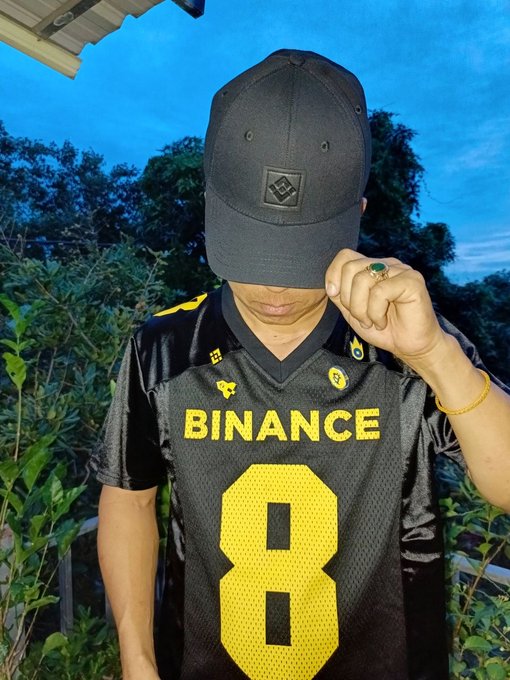 A person wearing a black cap and a black and yellow jersey with "BINANCE" and the number "8" printed on it stands outdoors with foliage in the background.