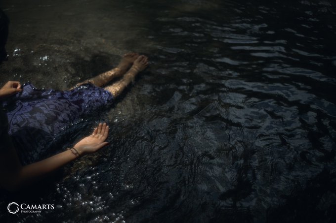 A person in a dark purple patterned dress lies at the edge of shallow, rippling water, with one hand reaching out toward the surface, photographed by Camarts Photography.