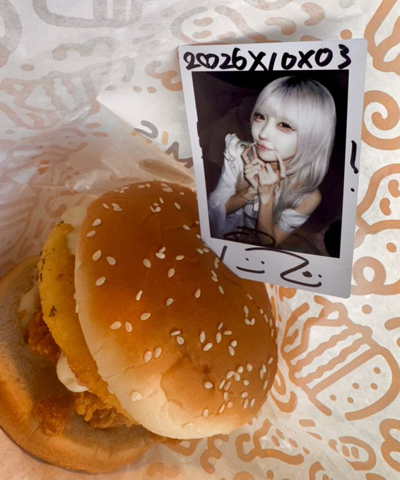 A polaroid-style photo of a person with platinum blonde hair and pink makeup making a heart gesture, placed next to a sesame seed bun burger on branded wrapper paper; the photo has text 'FOTOBOX 2002' on it.