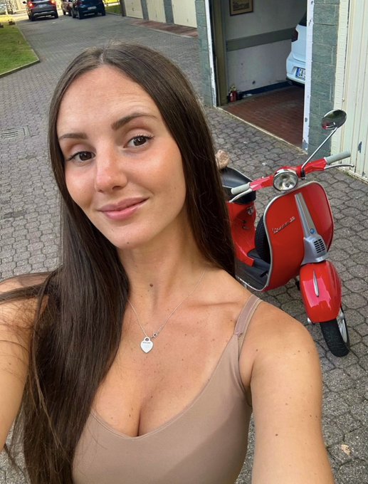 A young woman with long brown hair smiles for a selfie in front of a red Vespa scooter parked outside a building. The "Happy weekend :)" caption fits the relaxed, cheerful vibe of her weekend outing.
