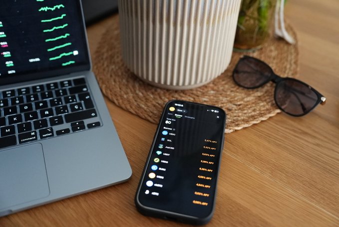 A smartphone on a wooden desk displays cryptocurrency prices including APX, BONK, USDC, JUP, and Pnut in a Jupiter mobile app interface, alongside a laptop showing green trading charts. The image illustrates the "borrow against your assets" concept by showing a portfolio on the @jup_mobile app that could be used as collateral for instant loans.