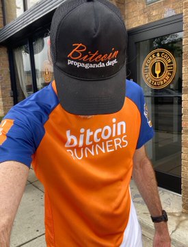A person wearing a black "Bitcoin propaganda dept." cap and an orange and blue "bitcoin RUNNERS" athletic shirt stands outside a building with a "BEER & COFFEE COMPANY" sign in the window.