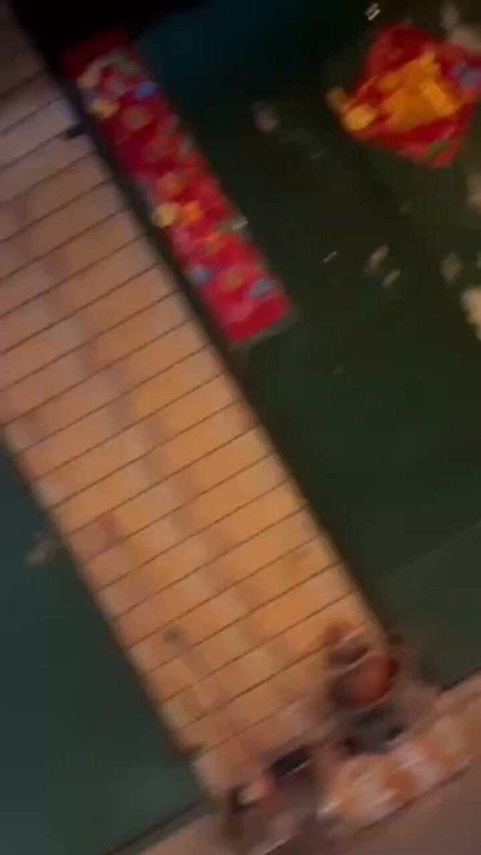 The image shows a person taking a picture of an item, possibly a toy or decoration, with their phone. The photo appears to be taken from a low angle, making the object appear larger than life. The background features a green wall adorned with various decorations, including a red banner and a yellow and orange striped sign. The person's action of capturing this scene suggests that they are interested in sharing it on social media or with friends.