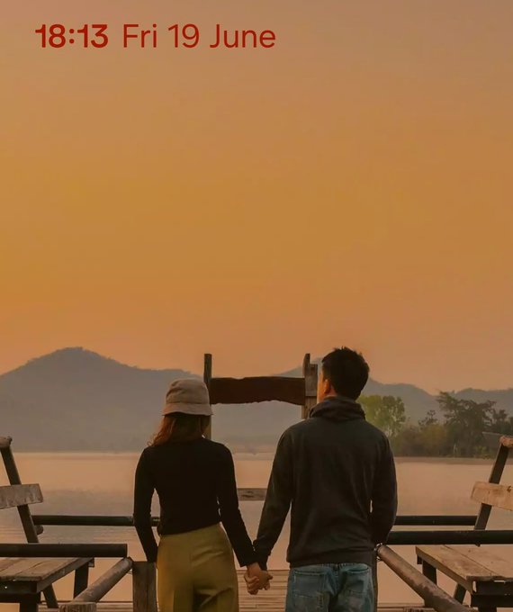 The image shows two people standing together, holding hands while looking out over a body of water at sunset. The couple appears to be enjoying each other's company as they gaze into the distance. In the background, there are benches where others might sit and watch the beautiful view. The setting sun casts an orange glow on the scene, creating a warm and romantic atmosphere.