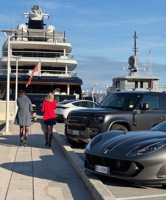 A street scene in a harbor featuring a large dark blue superyacht docked alongside a smaller gray naval vessel, with two women in coats walking past parked luxury cars including a gray Ferrari and a black Land Rover Defender with license plate 'GL 498 MG'.