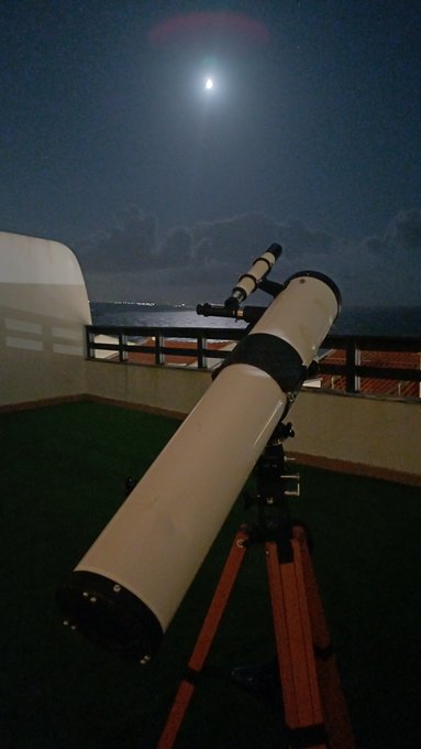 A telescope on a tripod points toward the night sky, where a bright full moon illuminates clouds over the ocean, viewed from a balcony.