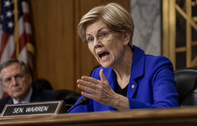 Senator Elizabeth Warren, identified by a nameplate, speaks and gestures emphatically while seated at a desk in a wood-paneled hearing room.