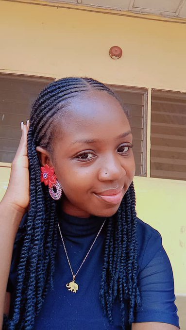A young woman with long black braided hair poses for a close-up selfie, wearing a navy blue top, red flower-shaped earrings, and an elephant pendant necklace, in front of a yellow building with brown windows.