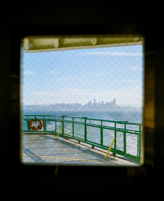 A view from a ferry or boat deck looking out toward a city skyline, with a green railing and a red and white life ring visible through a window frame.
