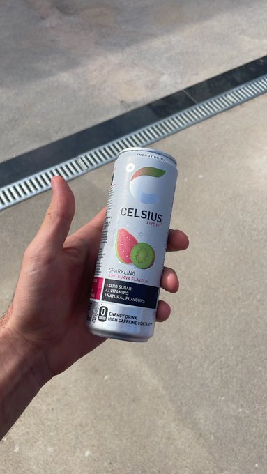 A hand holding a can of CELSIUS Sparkling Guava Flavour energy drink outdoors near a drainage grate.