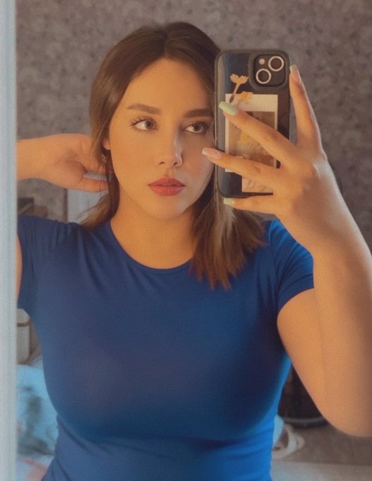 A woman taking a mirror selfie with her iPhone, wearing a blue t-shirt, with light green/pastel nails.