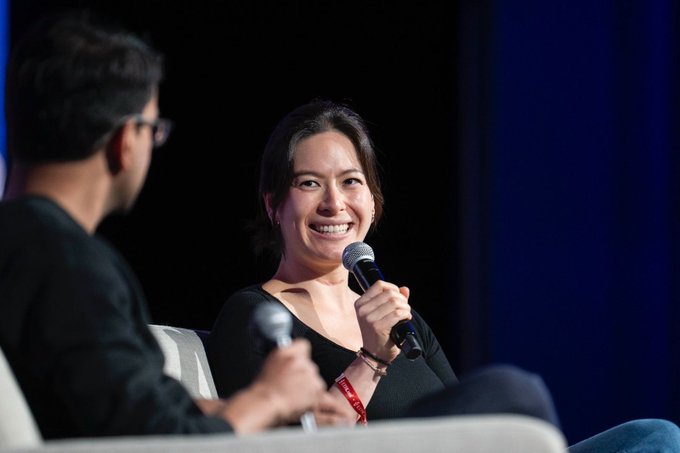 The image shows a man and woman sitting next to each other on a couch or chair, both holding microphones as they appear to be speaking into them. They are positioned close together, with the woman's microphone being held higher than the man's. The background is dark blue, providing a contrast that makes their interaction more prominent in the image.