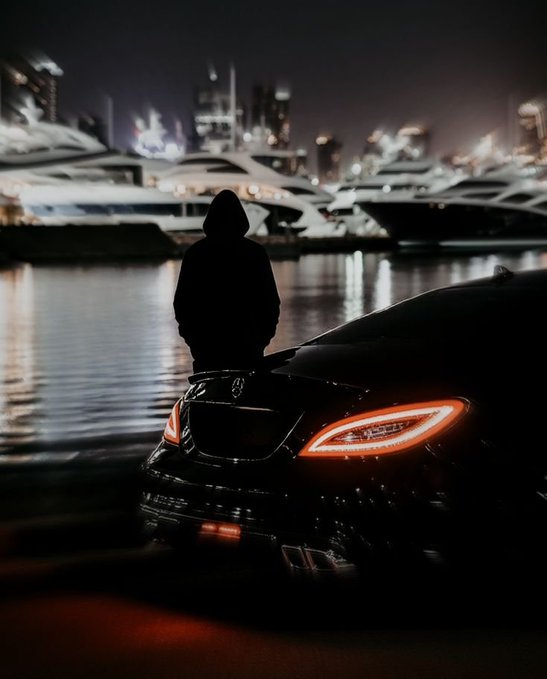 A hooded figure in silhouette leans against a dark car with illuminated red taillights, overlooking a marina full of luxury yachts at night with a city skyline in the background.