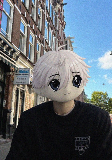 A person stands on a European-style street wearing a black Tommy Jeans sweatshirt, with a large anime-style plush head featuring white hair and big blue eyes covering their actual head.