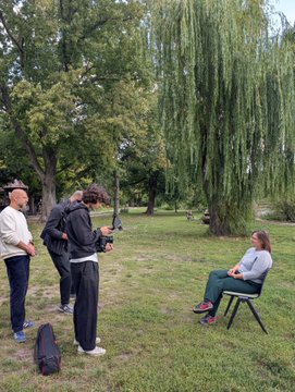An outdoor park scene where a film crew is conducting an interview; one person operates a camera on a gimbal while a woman sits on a chair being interviewed, with another man standing nearby.