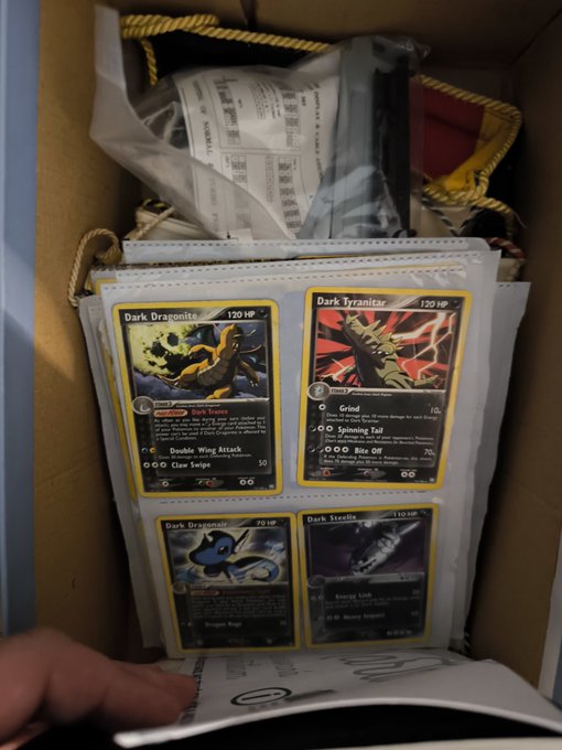 In the image, a person's hand is holding a card with Pokemon cards inside of an open cardboard box. The person appears to be looking at or inspecting the contents of one of these Pokemon decks as they hold up another card from their hand. This suggests that they might have found some Pokemon cards in the box and are examining them further.