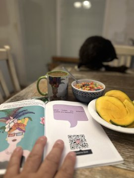 A cozy scene shows a person reading an open book at a wooden table, accompanied by a green mug, a bowl of cereal, and a plate with sliced mango and banana, while a black cat rests in the background.