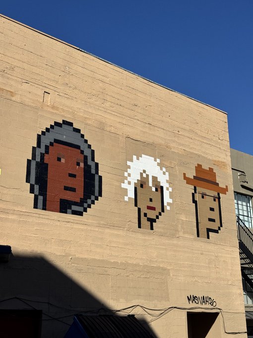 A pixel-art style mural painted on a beige brick wall depicts three stylized faces: a person in a dark hood, an older woman with white hair, and a man wearing a brown cowboy hat, signed 'MBU VANCO' at the bottom right.