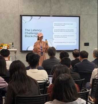A presentation slide titled 'The Latency Challenge Blockch...' being delivered to an audience by a person wearing a hijab at a conference.