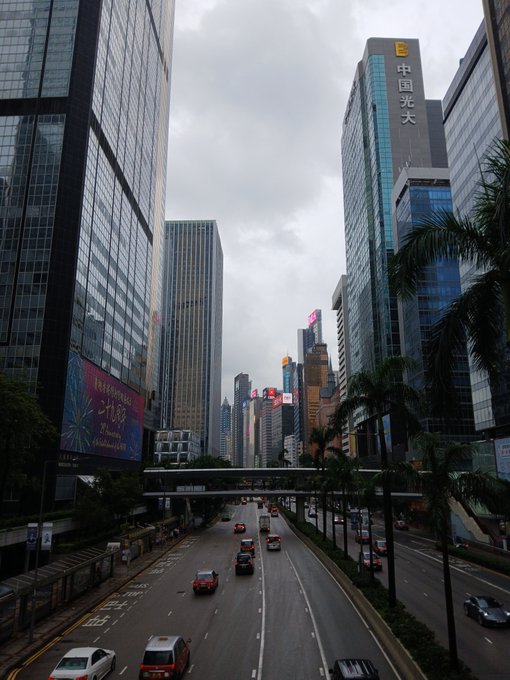 A street-level view of a Hong Kong CBD road flanked by tall skyscrapers, including one labeled '中国光大' (China Everbright), under an overcast sky.