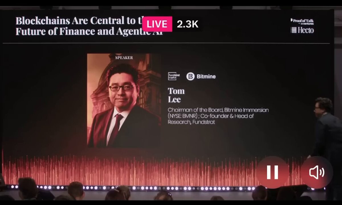 The image shows a large screen with a presentation slide featuring an image of a man wearing glasses, possibly Tom Lee, who is talking about cryptocurrency. The background of the image has red lines that appear to be part of a sound wave or music track. There are also some people in the scene, but they are not as prominent as the main focus on the presentation. The text at the bottom of the screen reads "Blokchains Are Central To i) Blockchain of Finance and Agencies Future of Finance and Agencies".