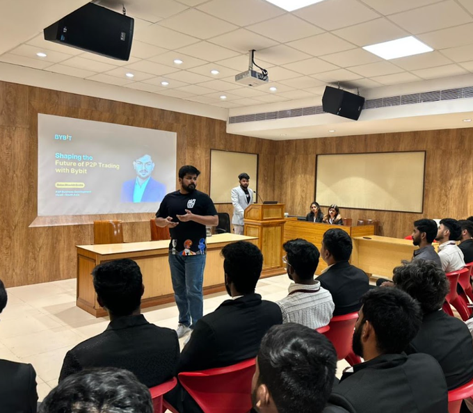 A man in a black t-shirt giving a presentation at a Bybit event titled 'Shaping the Future of P2P Trading' to an audience seated in a wood-paneled hall.
