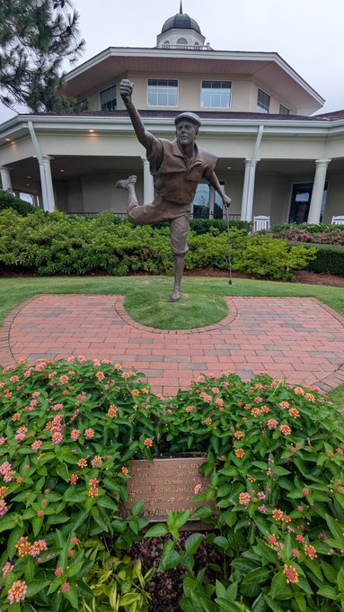 A bronze statue of a golfer mid-swing, set on a circular brick platform in front of a clubhouse with a domed tower, surrounded by green landscaping and flowers.
