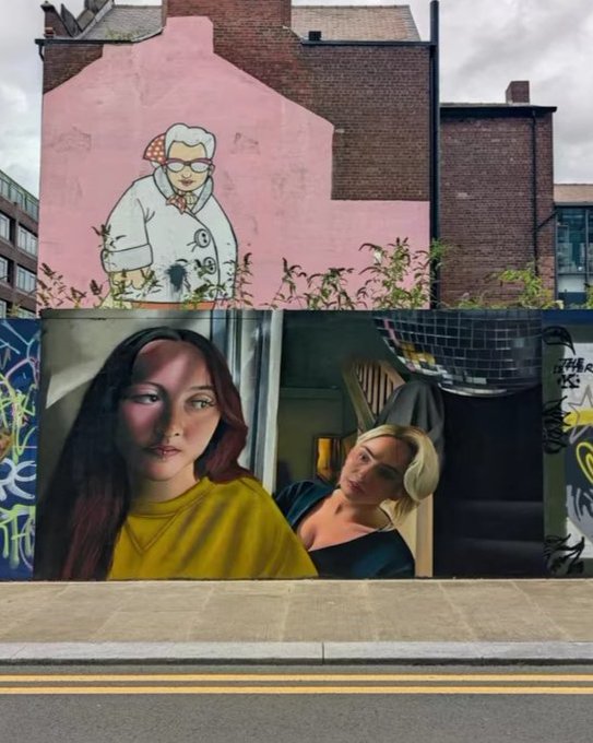A street view featuring two large murals on a building wall: an upper cartoon-style painting of an elderly woman on a pink background, and a lower realistic mural of two women.