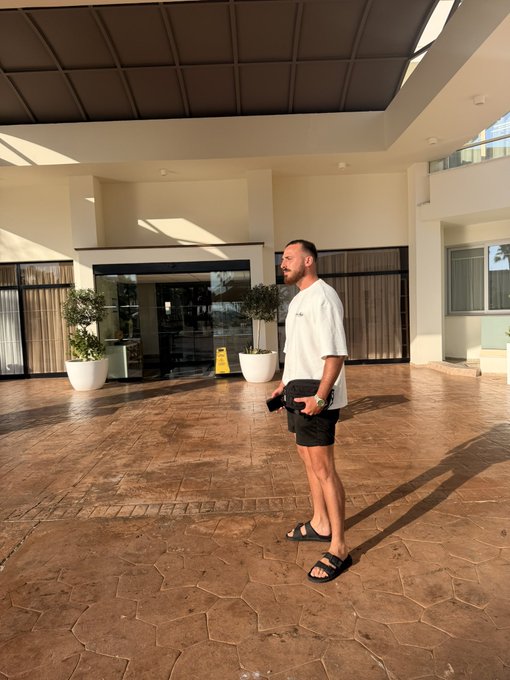 A man wearing a white t-shirt and black shorts stands on a stone patio at what appears to be a hotel entrance, holding his phone and smiling as sunlight casts long shadows across the scene—a perfect relaxed "Gm friends" morning greeting.