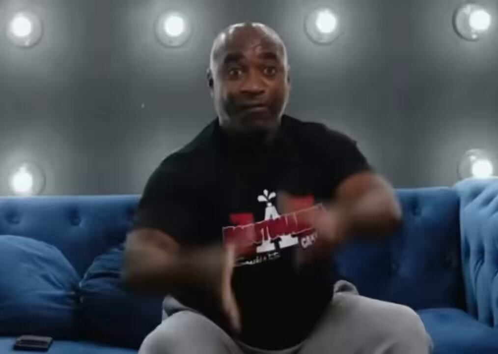 The image features a man sitting on a couch, wearing a black t-shirt with red text that says "I'm not a dumbass", and making a funny face as if he's being heckled by someone. The background of the image is dark gray, which contrasts with the bright white lights in the room. There are also two blue pillows on the couch behind the man.