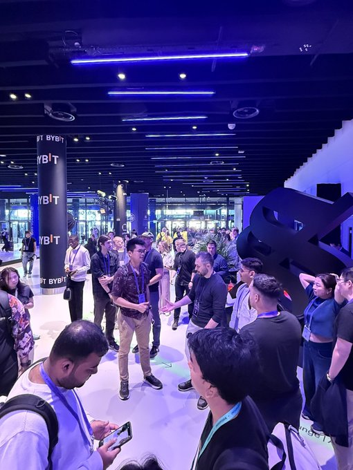 A crowded indoor event space bathed in blue lighting, with numerous attendees gathered around; branding for 'BYBIT' is visible on pillars in the background.
