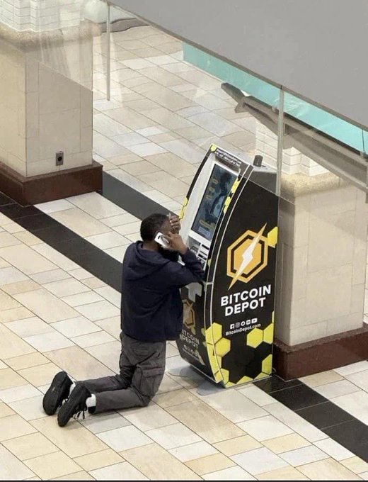 A man kneels and presses his phone to his ear while using a "BITCOIN DEPOT" ATM, appearing to plead with the machine in a mall setting. The image humorously illustrates the desperation of trying to acquire more $DOG cryptocurrency on Bitcoin, with the man's posture suggesting he's begging the ATM for just one more transaction.