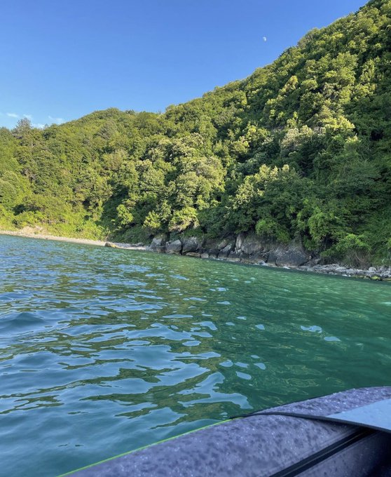 A serene view from a boat of turquoise waters lapping against a lush, forested shoreline on a bright sunny morning, perfectly matching the peaceful "good morning" sentiment of wishing friends a great day ahead.