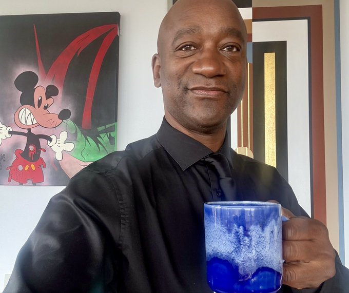 A smiling bald man wearing a black shirt and tie holds a blue ceramic mug in front of a wall featuring a colorful Mickey Mouse painting and framed artwork.
