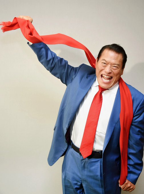 A man in a blue suit yells enthusiastically while swinging a red tie overhead against a plain wall.