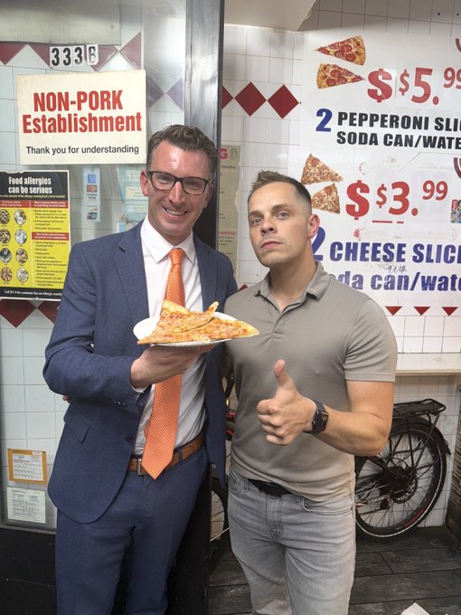 Two men pose in front of a pizzeria; one wears a suit and holds a slice of pizza, while the other in a polo shirt gives a thumbs up next to signs reading 'NON-PORK Establishment' and '2 PEPPERONI SLICES $5.99'.