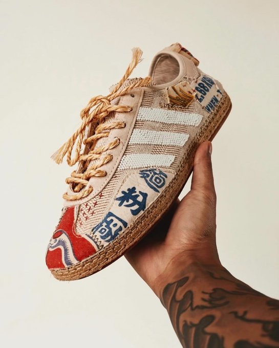 A hand holds a beige Adidas Gazelle-style sneaker featuring rope laces, woven textile panels with Asian characters, red suede accents, and an espadrille-style rope sole.