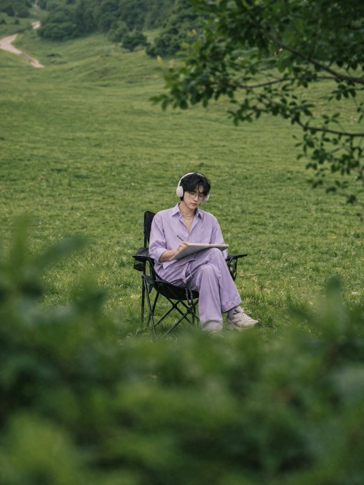 A photograph of a person wearing headphones and a lavender outfit, sitting in a chair on a green grassy hillside while drawing or writing in a sketchbook.