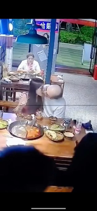 A man and woman are kissing while seated at a restaurant table with hot pot food, with another woman eating in the background and a sign displaying '5月' (May) visible.