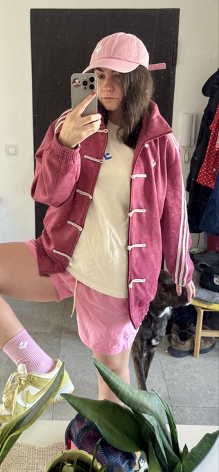 A mirror selfie of a young woman wearing a pink baseball cap, a maroon Adidas track jacket, a white t-shirt, and pink shorts, with her dog visible in the background.
