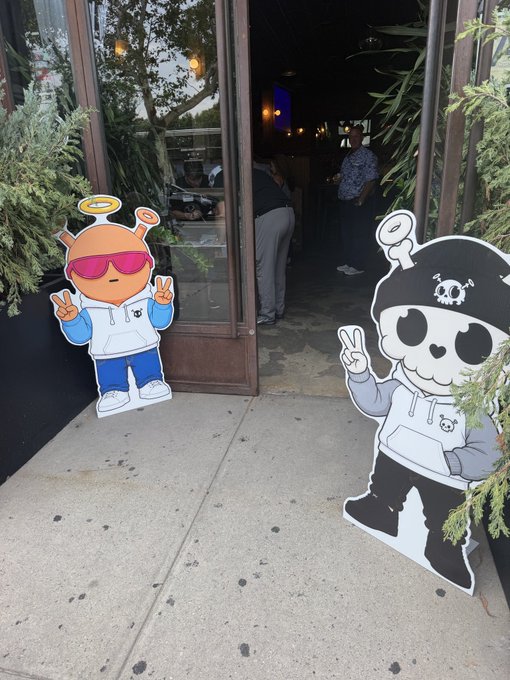 Two cardboard cutouts of cartoon characters—one orange and one a skeleton in a hoodie—are placed outside the entrance of a venue.