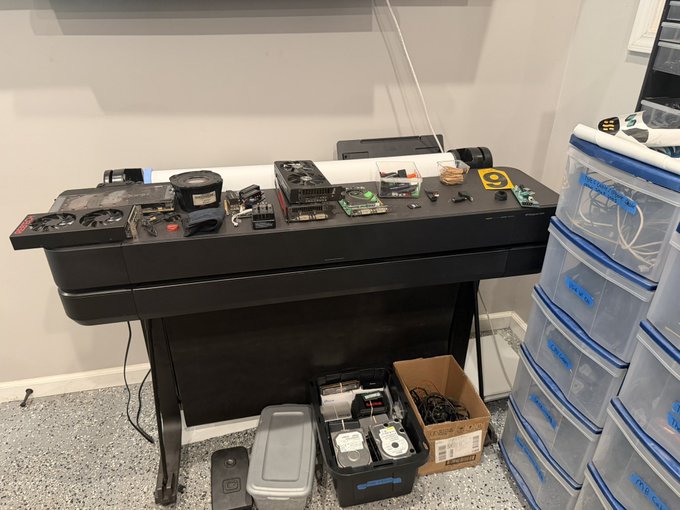 A black desk holds computer hardware components, including graphics cards and circuit boards, alongside a stack of clear plastic storage drawers labeled with 'X16' and other mining rig identifiers.