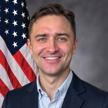 A professional headshot of a smiling man with light brown hair, blue eyes, and a beard, wearing a dark blazer over a light blue shirt, posed in front of an American flag.