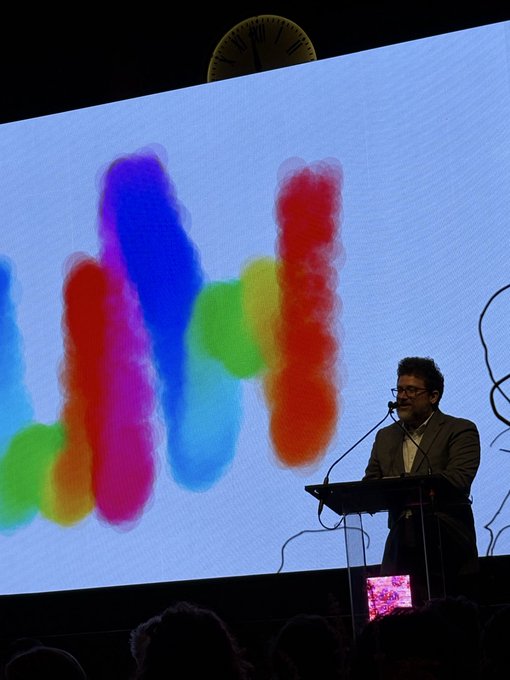A man with glasses speaks at a podium on a stage, standing in front of a large screen displaying colorful abstract artwork.
