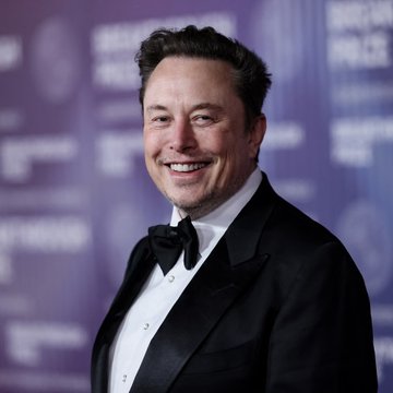A smiling man in a black tuxedo with a bow tie stands in front of a purple backdrop featuring partially visible text including "MUSK." The image relates to the post about Elon Musk's bold claim regarding SpaceX's AI development timeline.