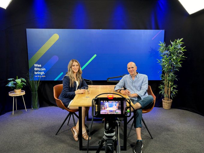 A studio interview setting with a woman and a bald man seated at a wooden table, with a large screen behind them displaying 'Was Bitcoin bringt,' suggesting a German-language show about Bitcoin.