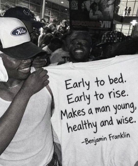 A black and white photo shows a smiling man wearing an 'Hus' trucker hat while holding up a white t-shirt printed with the Benjamin Franklin quote 'Early to bed. Early to rise. Makes a man young, healthy and wise.'