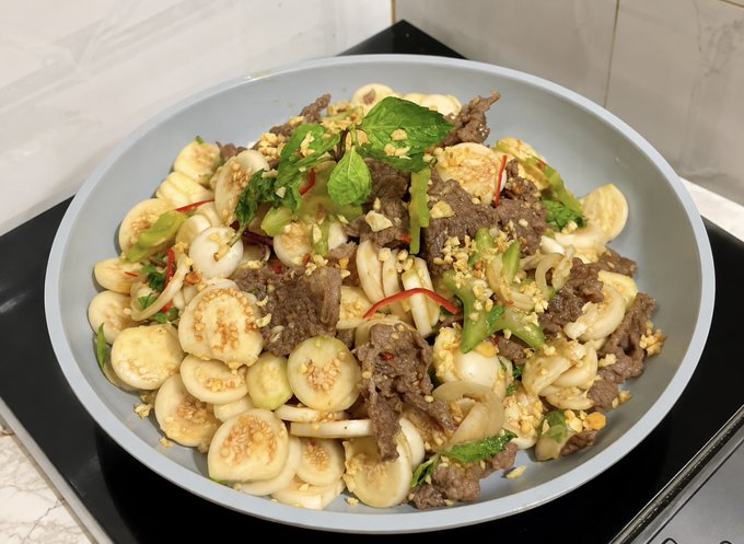 A plate of stir-fried beef with sliced eggplant, onions, green peppers, basil leaves, and chili peppers, garnished with crushed peanuts and served on a light gray dish.