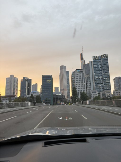 A view from inside a car driving across a bridge toward a city skyline with tall skyscrapers, including one with 'MAN' visible on top, under a hazy sunset sky.