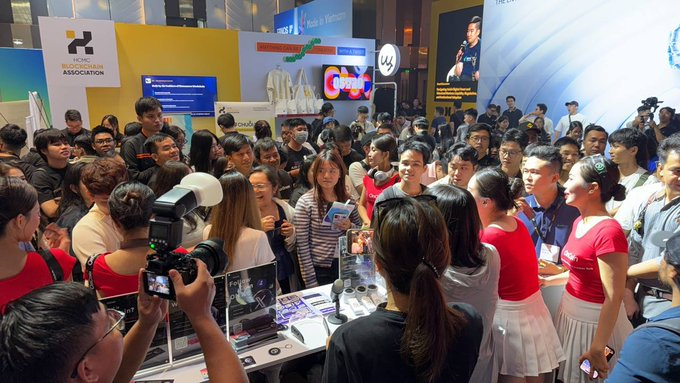A crowded convention booth features signage for the "HCSC Blockchain Association" as attendees gather to pick up "Oncoin" merchandise, with a photographer capturing the lively scene around booth #20.